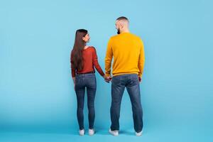 A young married couple stands together, holding hands and looking at each other affectionately. They wear casual outfits, looking relaxed and happy against a bright blue backdrop, back view photo