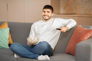 A young Caucasian man lounges comfortably on a sofa, holding a remote and a bowl of popcorn. He is dressed casually and appears relaxed, enjoying a leisurely afternoon indoors. photo