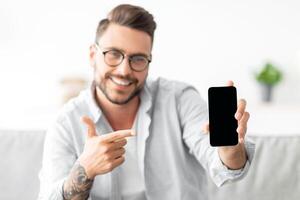 Smartphone mockup. Millennial man showing cellphone with blank screen and pointing finger on it, space for your ad or design. Guy sitting on sofa at home, holding phone in hand photo