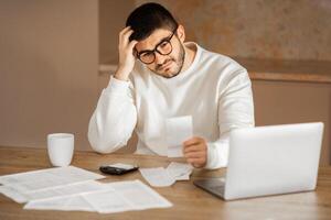 A young European man wearing eyeglasses sits at a wooden table in his home, looking puzzled as he examines a document. photo