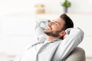Lazy day. Happy relaxed man resting on comfortable armchair, holding hands behind head, sitting with closed eyes and enjoying silence and comfort of minimalism lifestyle, free space photo