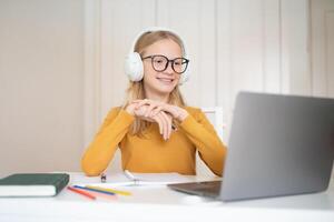 A European teenage girl with glasses enjoys online learning at home. She sits at a desk, smiling while engaged with her laptop, surrounded by school supplies and a notebook. photo