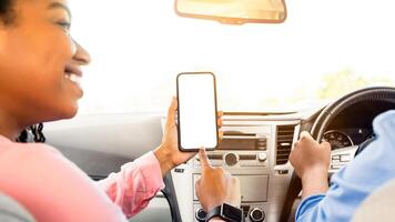 Look Here. Rear back view of happy African woman using cell, pointing at smart phone blank white screen inside a car, searching location via gps navigator application, man driving automobile, close up photo