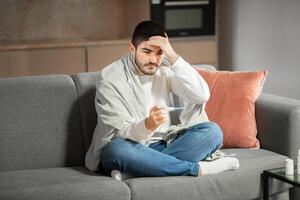 A young European man in eyeglasses sits on a couch at home, wrapped in a blanket. He appears concerned as he checks his temperature with a thermometer, showing signs of illness. photo