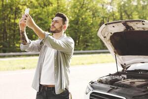 Young guy standing near his broken car after auto accident, looking for mobile network signal, trying to call roadside assistance, having no connection, needing help in countryside photo