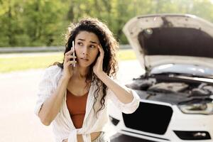 Young woman with smartphone calling towing service on road, having phone conversation with emergency service near her broken auto on highway. Road accident, car wreck, engine failure concept photo
