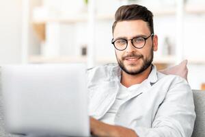 Handsome guy with glasses using laptop at home, sitting on sofa in living room, free space. Happy man freelancer working from home, typing emails or programming photo