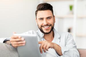 Handsome young man holding digital tablet and smiling at camera, browsing internet or checking social networks, sitting on couch at home, free space. Happy guy using modern technologies for leisure photo