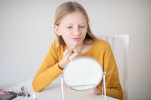 A teenage girl sits at a table in a bright room, carefully applying lip gloss while looking into a small mirror. Various makeup products are spread out on the table, showcasing her interest in beauty. photo