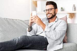 Great mobile app. Happy guy using smartphone, texting and surfing social networks, sitting on couch at home, copy space. Millennial man browsing internet, using app on cellphone photo