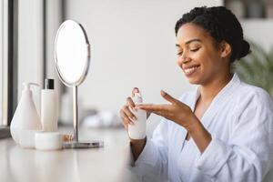 A woman smiles while applying a skincare product at home. She is seated near a window with natural light illuminating the space, showcasing her daily beauty routine in a relaxed setting. photo