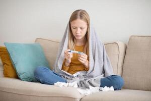 A teenage girl sits on the couch at home, wrapped in a blanket, looking concerned as she checks her temperature. Used tissues are scattered around, indicating she is unwell. photo