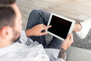 Young man using digital tablet with blank black screen, browsing website or showing new app, sitting on sofa at home. Advertisement concept. Device with empty screen, mockup for your design photo