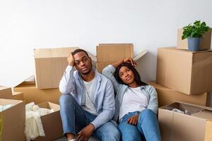Sad black spouses exhausted after moving to new apartment, sitting among cardboard boxes and resting, having problems during relocation to own home photo