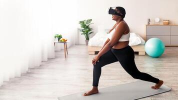 Side view of overweight black lady in VR headset training with virtual reality app, making lunges at home. Plus size Afro woman using modern technologies for online domestic training, full length photo