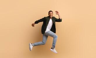A joyful African American man is leaping in a dynamic pose. He wears casual attire with jeans and a shirt, showcasing an upbeat lifestyle while posed against a soft, beige backdrop. photo