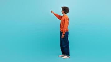 A young African American man stands with curly hair and an orange sweatshirt, pointing upward with enthusiasm in a bright, isolated setting, touching invisible screen photo