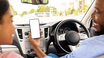 Back rear over the shoulder view of happy black male driver holding cell in hand showing white blank mock up screen to female passenger sitting on front seat using mobile app for city navigation photo