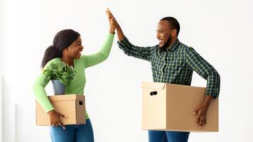 Happy Moving Day. Joyful Black Spouses Holding Cardboard Boxes And Giving High Five To Each Other, Cheerful African American Couple Celebrating Relocation To Their Own Apartment, Copy Space photo