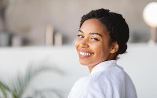 African American woman with curly hair smiles brightly while wearing a cozy spa robe. She seems relaxed and content in a calm and soothing setting, radiating happiness and tranquility, copy space photo