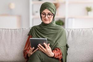 Senior Muslim Female Using Digital Tablet Reading Book Online Sitting On Couch At Home. Modern Middle-Eastern Housewife In Hijab And Glasses Watching Movie, Browsing Internet Via Computer photo