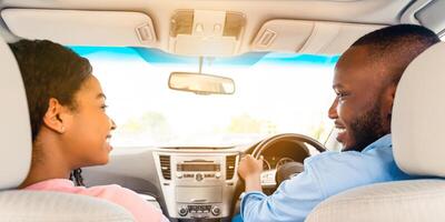 Rear back view of smiling handsome African American man driving modern luxury car, happy beautiful young woman sitting on the front passenger seat, looking at each other, sun flare photo