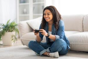 A young woman sits comfortably on the floor, playing games with a joyful expression. The cozy living room features a modern sofa and indoor plants, creating a relaxing atmosphere. photo