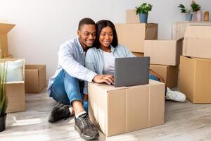 Happy african american spouses having call via laptop, sitting among carton boxes in new apartment and smiling to computer webcamera. Black couple telling news about relocation to family photo