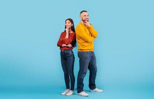 A young couple poses playfully back to back against a blue background, showcasing their affection. They exhibit thoughtful expressions while enjoying each other's company. photo