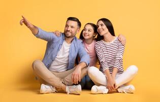 A father, mother, and their daughter sit together on the floor, showing affection. The father points excitedly, while the mother and daughter share smiles. photo