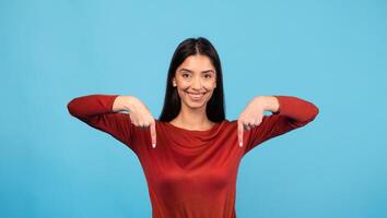 A millennial woman with brunette hair smiles brightly while playfully pointing downward. She is isolated against a solid blue background, showcasing a lively expression in a studio setting. photo