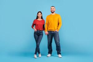 A millennial couple stands together, holding hands and smiling. The woman wears a red sweater while the man sports a yellow sweater, both exuding warmth and affection in a cheerful setting. photo