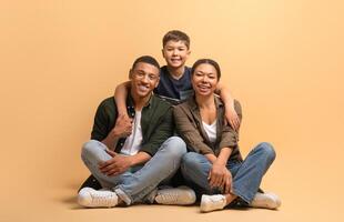 A joyful family of three shares a warm moment with smiles. The father and mother sit casually with their son, enjoying their time together against a bright background. photo