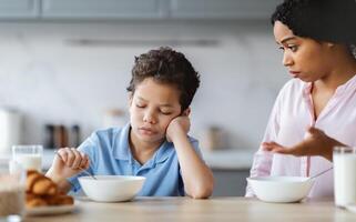 A mother talks to her son in a kitchen while he eats breakfast. The child appears thoughtful and a bit sad as the parent expresses concern and love, showcasing family dynamics. photo