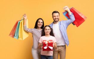 A cheerful family stands together against a bright yellow background, celebrating their shopping trip with colorful bags. The daughter holds a beautifully wrapped gift, showcasing their happiness. photo