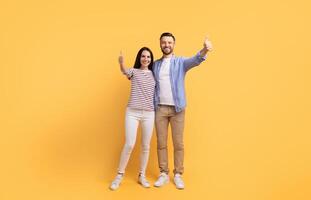 A cheerful couple stands together in front of a vibrant yellow background, both giving a thumbs up. They share a moment of happiness and love, showcasing their strong connection. photo