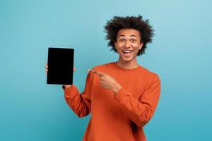 A cheerful young man with curly hair is pointing at a tablet in a vibrant blue environment. He wears an orange long-sleeve shirt and has an expressive smile, embodying a positive lifestyle. photo