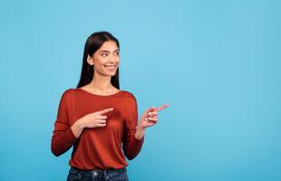 A millennial woman with brunette hair stands against a vibrant blue backdrop, smiling and pointing with both hands at copy space. Her expression conveys excitement and positivity in a casual setting. photo