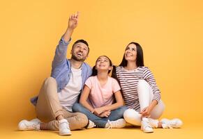 A happy family smiles while sitting cross-legged on the floor. The mother and father point at something above, engaging with their daughter, who looks excited and joyful. photo