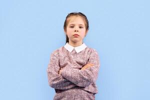 A girl stands confidently with her arms crossed, displaying a cheerful expression. Her happy demeanor contrasts with the simplicity of the blue backdrop, creating a vibrant scene. photo