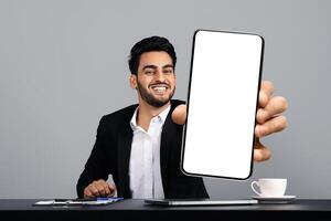 A young Arab entrepreneur sits at a desk in a modern studio, smiling confidently as he holds up a blank smartphone screen. He is dressed in a formal black suit, exuding a successful lifestyle. photo