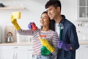 Cute asian couple taking selfie while cleaning kitchen together, using sponge as smartphone, hugging and smiling, young man and woman having fun while house-keeping at home, side view photo