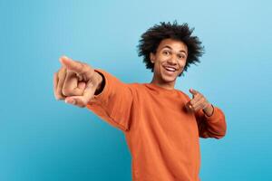 A cheerful African American man in a casual orange sweater smiles widely while pointing playfully at the viewer. The vibrant blue background enhances his lively demeanor. photo