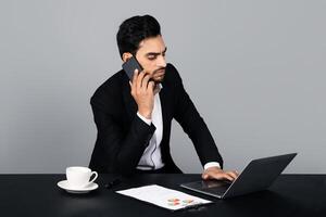A Middle Eastern businessman is focused on a phone call while typing on his laptop in a modern studio. photo