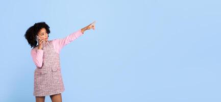 A joyful preteen girl engages in a phone conversation, smiling and pointing at copy space as if sharing an exciting story. She stands alone against a bright blue backdrop photo