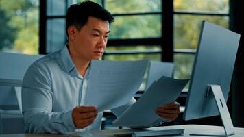 Tired unhappy Asian man worker manager sit at office doing routine paperwork read contract documents budget invoice pay bills online analyze business plan check documentation professional paper audit photo