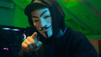 Lviv, Ukraine - June 03, 2024. Hacker anonymous typing computer looking at camera pointing finger programmer attack cyberspace fraud burglar internet spyware virus pc scam network binary code firewall photo