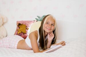 Cute Caucasian teen girl reads her diary sitting on the bed in sleeping clothes and makes notes in it photo