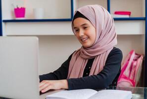 Portrait of a happy female Muslim student in hijab sitting at a desk using a laptop computer and learning for school in the classroom or at home. photo