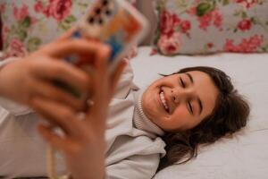 A smiling teenage girl using a phone while comfortable lying on the bed at home. photo
